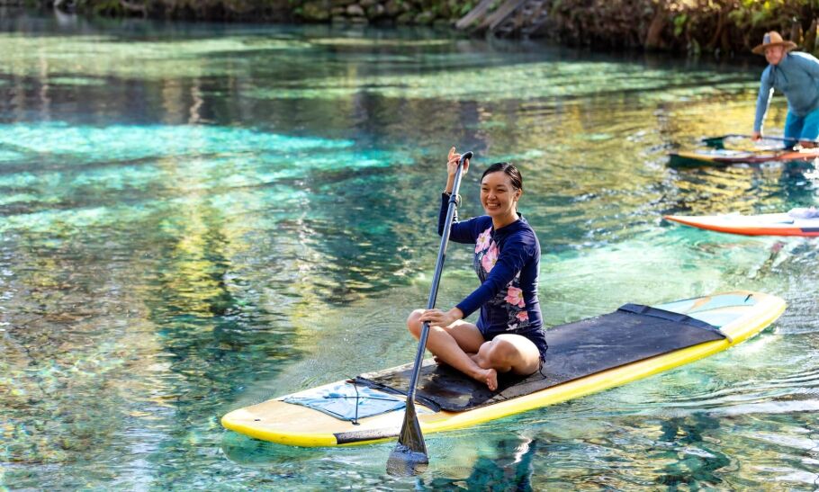 An Asian American woman sitting crossed-legged on a stand up paddleboard. She is paddleboarding on a tranquil Florida river enjoying nature. She is smiling and looking at the camera.